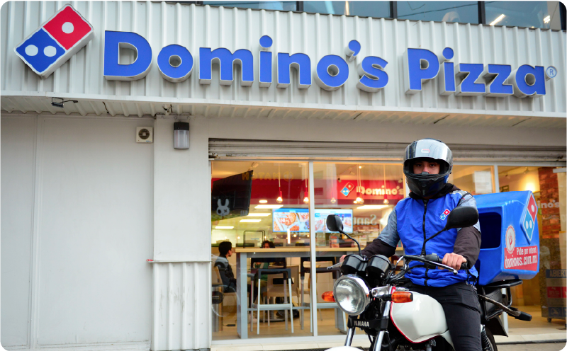 Delivery person on a motorcycle in front of a Domino's Pizza store, wearing a blue Domino's jacket and helmet, ready to deliver pizzas