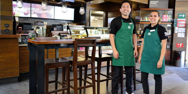 A group of five Starbucks baristas in green aprons standing together in a store, smiling and waving at the camera. Some are wearing masks while others are showing peace signs with their hands.