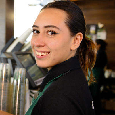 A smiling Starbucks barista in a green apron and black cap holding up a frappuccino drink in a clear plastic cup with whipped cream on top, standing in front of a Starbucks counter with menu displays visible in the background.