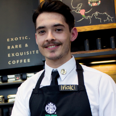 A female Starbucks barista with a white cap and green apron standing confidently with arms crossed in a Starbucks store with a warm smile on her face.