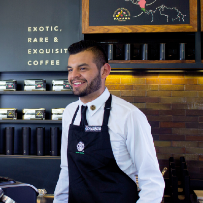 A male Starbucks barista wearing a black mask, black cap and green apron holding a pen, ready to take an order or write on a cup in a Starbucks store with brick wall background.