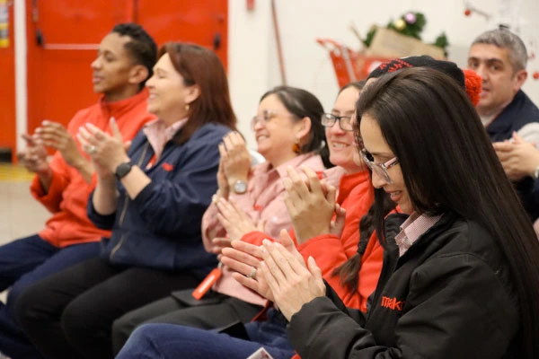 Un grupo de empleados diversos de Makro sentados en fila en un evento de la empresa, vistiendo ropa corporativa roja y azul marino. Todos están sonriendo y aplaudiendo con entusiasmo. La imagen captura un momento de unidad de equipo y cultura corporativa positiva en un entorno interior brillante con acentos rojos que coinciden con los colores de la empresa.